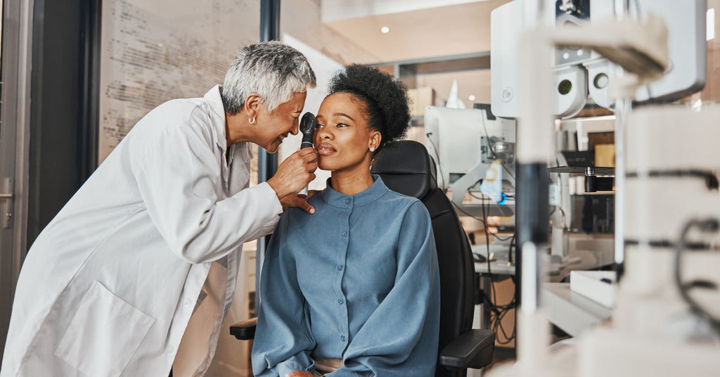 A woman sitting in a chair while a doctor examines her right eye. They are both in a doctor’s office.