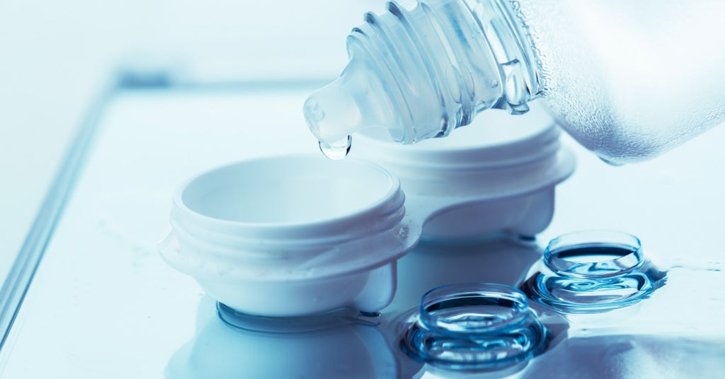 A bottle of contact lens solution is above a lens case and a drop of solution is hanging onto the edge of the dropper.