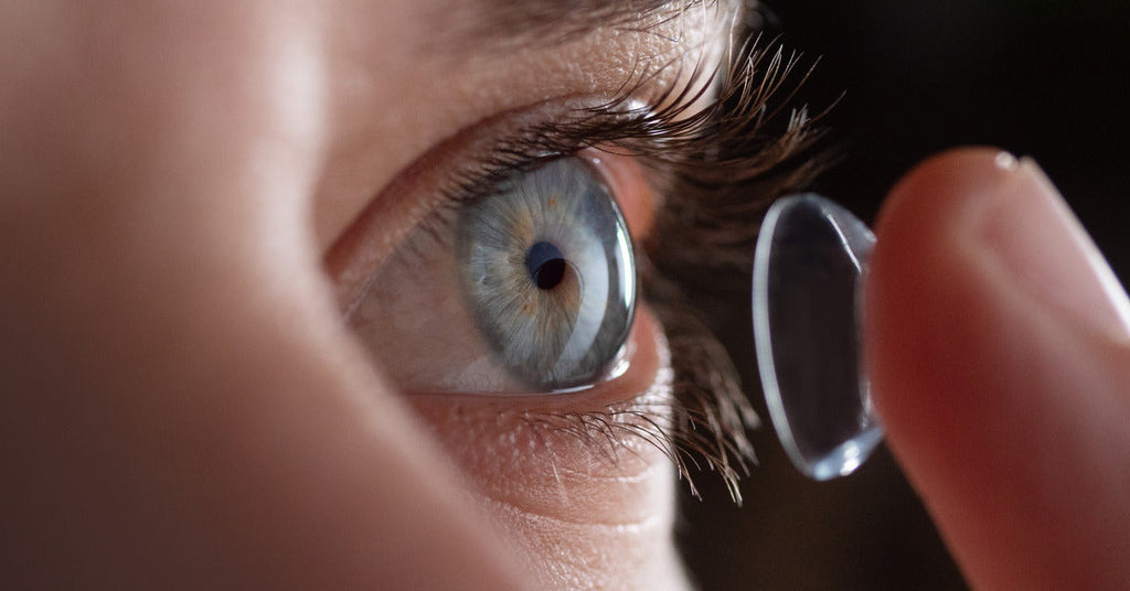 A person in front of a black background holding a contact lens on their fingertip before placing it in their eye.