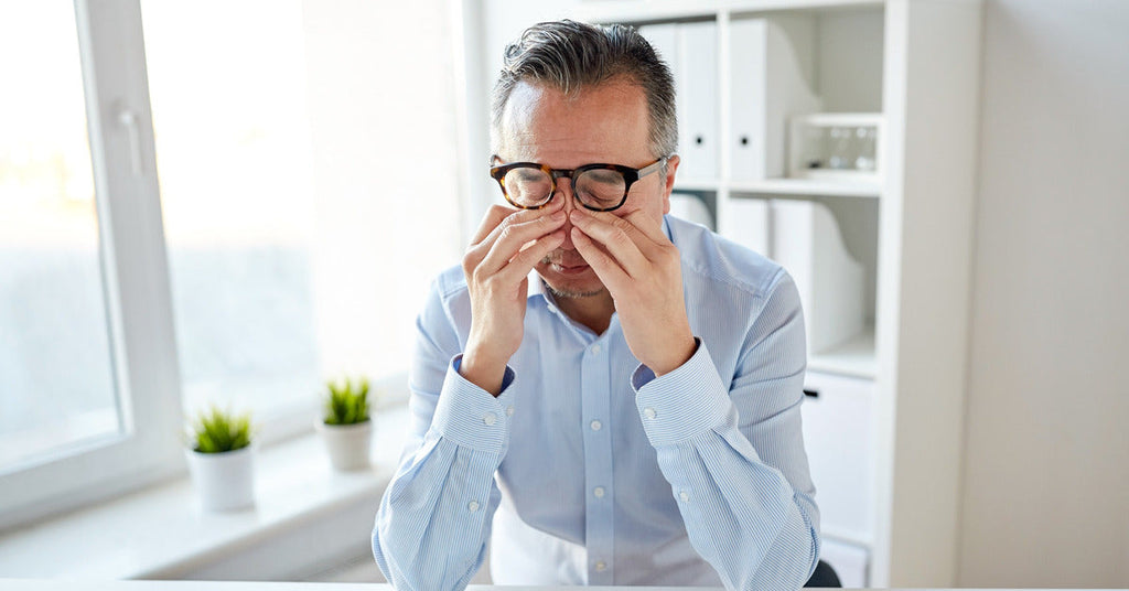 An office worker leans over a desk and rubs their eyes. The room is mainly white and dull, except for two green plants.