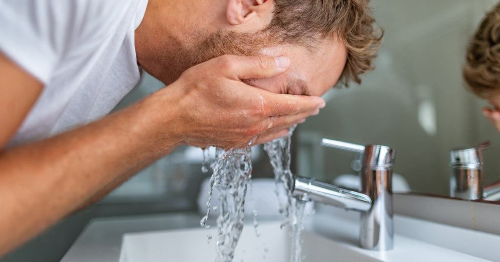 A person wearing a white shirt leaning over a sink and splashing water in their face. A mirror is in front of the sink.