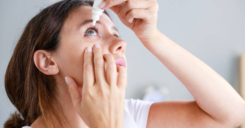 A woman in a white t-shirt looks up while she holds a white bottle of eye drops above her eye.